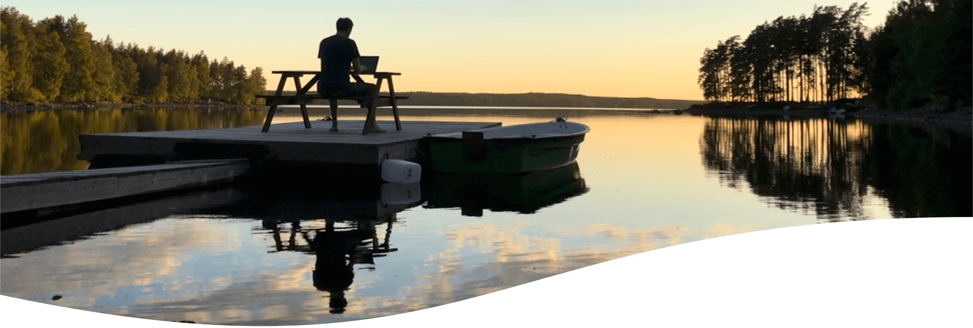 Philosoph Dr. Nicolas Dierks schreibt inmitten der Ruhe eines Sees in Schweden - er sitzt am Laptop auf einem Holzsteg, an dem ein kleines Ruderboot liegt. 